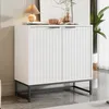 A white cabinet with fluted doors and black metal legs is placed in a room with a light gray floor and a beige wall. Produktbild-Vorschau