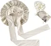 A beige satin bonnet with long ties is laid out on a white background. To the right, a small white bowl contains several white silkworm cocoons, with a vintage postcard underneath. Voir les images du produit