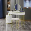 A white vanity table with gold accents and a round LED mirror, accompanied by a matching stool and a cylindrical drawer unit, set against a dark gray wall with a framed artwork. Náhled obrázku produktu