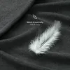 A close-up shot of a dark gray textured fabric, likely a bedsheet, with a white feather resting on it. Text overlays indicate softness and coziness. Anteprima dell’immagine prodotto