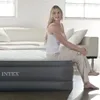 A woman is sitting on an air mattress in a room with a sofa and windows. Podgląd zdjęcia produktu