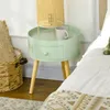 A mint green round side table with a drawer and wooden legs, next to a bed and a woven basket. Voir les images du produit