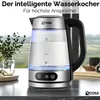 A glass electric kettle with a stainless steel base and handle is shown on a countertop, with a blurred background of greenery and a window. Náhľad obrázku produktu