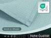 Close-up of a light blue waffle-patterned fabric, likely a blanket or throw, with certification labels and descriptive text overlays. Produktbild-Vorschau