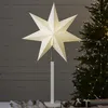 A white star-shaped lamp stands on a white base in front of a wooden wall and a Christmas tree with lights. Podgląd zdjęcia produktu