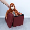 A hand is placing a stuffed orangutan and a stuffed dinosaur into a red storage ottoman. Náhled obrázku produktu