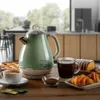A vintage-style green electric kettle is set on a wooden table with breakfast items like croissants, cake, and tea. Produktbild-Vorschau