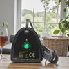 A black steam iron power station sits on a wooden table in front of a window with a view of greenery. A glass of amber liquid and a wicker basket with white laundry are also visible. Náhled obrázku produktu