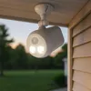 A white outdoor motion-activated spotlight is mounted under a wooden soffit, illuminating the exterior of a house. Produktbild-Vorschau
