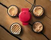 A red mini waffle maker with interchangeable plates is shown on a wooden table, surrounded by four finished waffles in different shapes: a pumpkin, a gingerbread man, a heart, and a classic round waffle. Produktbild-Vorschau