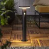 A black solar bollard light with a motion sensor is shown on a wooden deck at night, with a yellow chair and a large potted plant in the background. Podgląd zdjęcia produktu