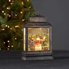 A decorative LED lantern with a Christmas scene inside, set against a blurred Christmas tree background. Podgląd zdjęcia produktu