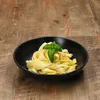 A black bowl of fettuccine pasta with a basil garnish sits on a wooden table. Podgląd zdjęcia produktu