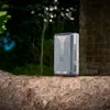 A battery pack sits on a tree branch outdoors, with trees and a brick wall in the background. Anteprima dell’immagine prodotto