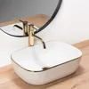 A close-up shot of a white oval countertop sink with a gold rim and a gold faucet, set on a wooden surface with a mirror in the background. Náhľad obrázku produktu