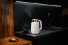 A modern kitchen counter with a steaming white kettle, a cup of coffee, and a croissant. Voir les images du produit