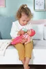 A young girl is feeding a baby doll with a bottle while sitting on a couch. Náhľad obrázku produktu