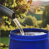 Water is flowing from a downspout into a blue barrel in an outdoor setting with greenery and trees in the background. Produktbild-Vorschau