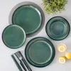 A top-down view of a set of green ceramic plates and bowls arranged on a light gray and white surface, with a small green plant in the upper right corner and lemons and a glass of water in the lower right corner. Voir les images du produit
