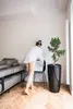 A woman is arranging a picture frame on a chair next to a large potted plant in a modern living room. Produktbild-Vorschau
