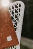 A brown blanket with a white tag is draped over a white wicker chair outdoors. Produktbild-Vorschau