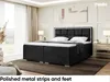A bedroom scene featuring a large black boxspring bed with a white mattress and topper, a black upholstered headboard with silver trim, and two bedside tables with lamps. The room has large windows with sheer curtains and a painting on the wall. Produktbild-Vorschau