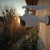 A security camera is mounted on a textured wall, with a garden and trees in the background during sunset. Produktbild-Vorschau