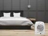 A white electric heater is placed on a wooden floor next to a bed in a modern bedroom. Produktbild-Vorschau