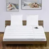 A bed with a mattress topper and pillows, with framed pictures on the wall above. Produktbild-Vorschau