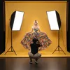 A photographer is taking a picture of a model in a studio with a yellow backdrop and studio lights. Náhled obrázku produktu