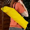 A person carrying a yellow bag over their shoulder, with a wooden fence and greenery in the background. Voir les images du produit