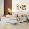 A bedroom scene featuring a tufted beige bed with pillows and blankets, a nightstand with a lamp and vase, and framed pictures on the wall. Produktbild-Vorschau