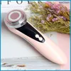 A pink facial massage device with silver and black accents is placed on a surface with a floral arrangement in the background. Náhľad obrázku produktu