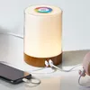 A bedside lamp with a wooden base and a frosted glass shade, connected to a smartphone and a small pink speaker via USB cables. Produktbild-Vorschau