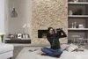 A woman is sitting on a rug in a living room, looking through a camcorder. The room has a stone fireplace and shelves with decorative items. Anteprima dell’immagine prodotto
