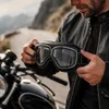 Mann hält ein Paar schwarze Vintage-Motorradbrillen mit Chromrahmen und klaren Gläsern, vor unscharfem Hintergrund Anteprima dell’immagine prodotto