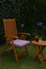 A wooden chair with a purple cushion and two drinks on a small wooden table in a garden setting. Produktbild-Vorschau