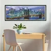 A framed puzzle of the New York City skyline at night is hanging on a wall above a wooden desk with a chair and a potted plant. Náhled obrázku produktu