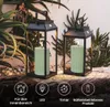 Two LED lanterns with green candles are placed on a wooden surface, with plants in the background. Podgląd zdjęcia produktu