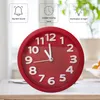 A red analog alarm clock with white numbers and hands, set on a table. The background shows a blurred indoor scene with plants and a window. Produktbild-Vorschau