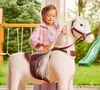 A young girl is brushing the mane of a white rocking horse in a backyard setting. Produktbild-Vorschau