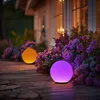 Two solar sphere lights, one purple and one orange, are placed on a wooden deck next to blooming purple flowers. The lights are illuminated, casting a soft glow. Produktbild-Vorschau