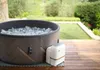An inflatable hot tub filled with water and bubbles sits on a wooden deck outdoors, with a control unit next to it. The background shows a manicured lawn and greenery. Náhled obrázku produktu