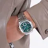 A close-up shot of a man's wrist wearing a silver watch with a green dial, partially covered by a tan jacket. Produktbild-Vorschau