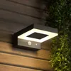 A modern black outdoor solar wall light with a square illuminated panel is mounted on a wooden wall, with green foliage hanging to the right. Produktbild-Vorschau