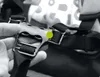 A hand is holding a black buckle mechanism with straps attached to it. A green dot is visible on the buckle. Produktbild-Vorschau