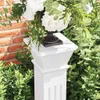 A close-up view of a white plant stand with a floral arrangement on top, set against a brick wall and lush green foliage. Anteprima dell’immagine prodotto