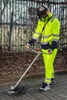 A person wearing high-visibility workwear is using a string trimmer on a paved area next to a fence and some foliage. Produktbild-Vorschau