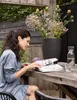 A woman sits at a wooden table outdoors, reading a magazine next to a large planter filled with flowers. A carafe of water and a glass are also on the table. Produktbild-Vorschau