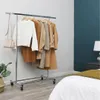 A garment rack with several coats and a sweater is displayed in a room with a bed in the foreground and a teal accent wall in the background. Náhled obrázku produktu